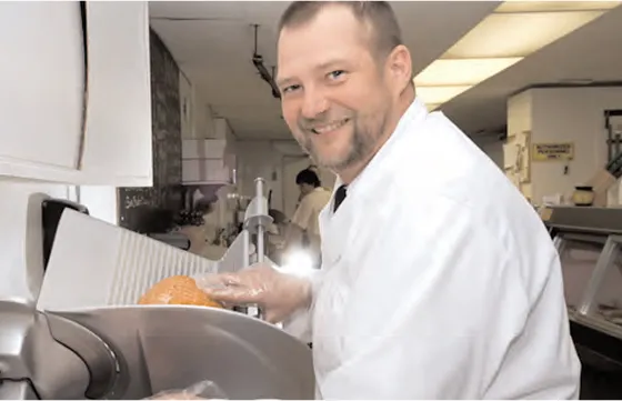 A smiling man in a white coat and gloves is slicing deli meat in a grocery store deli department.