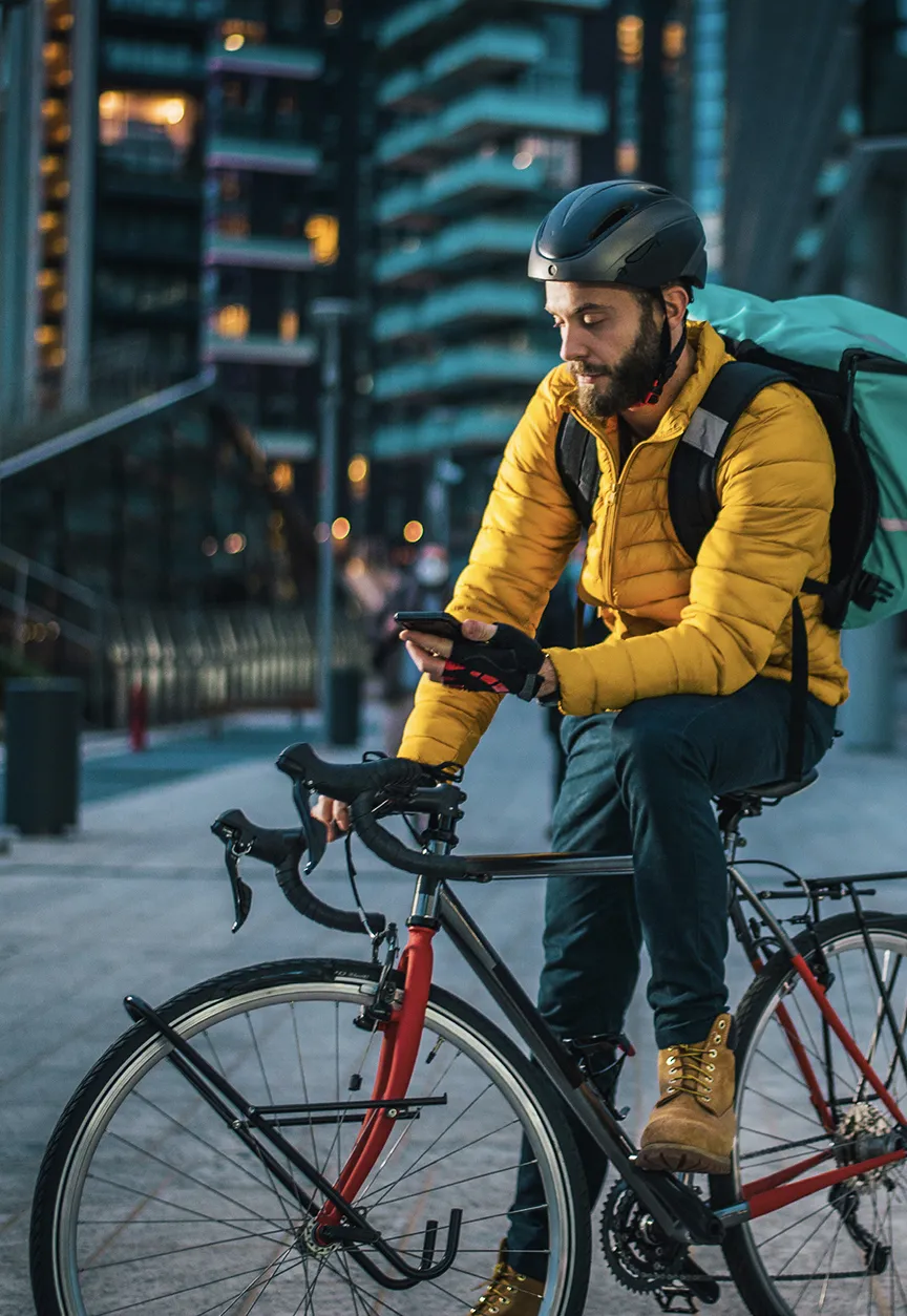 Food delivery worker wearing a jacket, helmet and delivery backpack sits on a bike checking his phone in an urban setting at dusk.