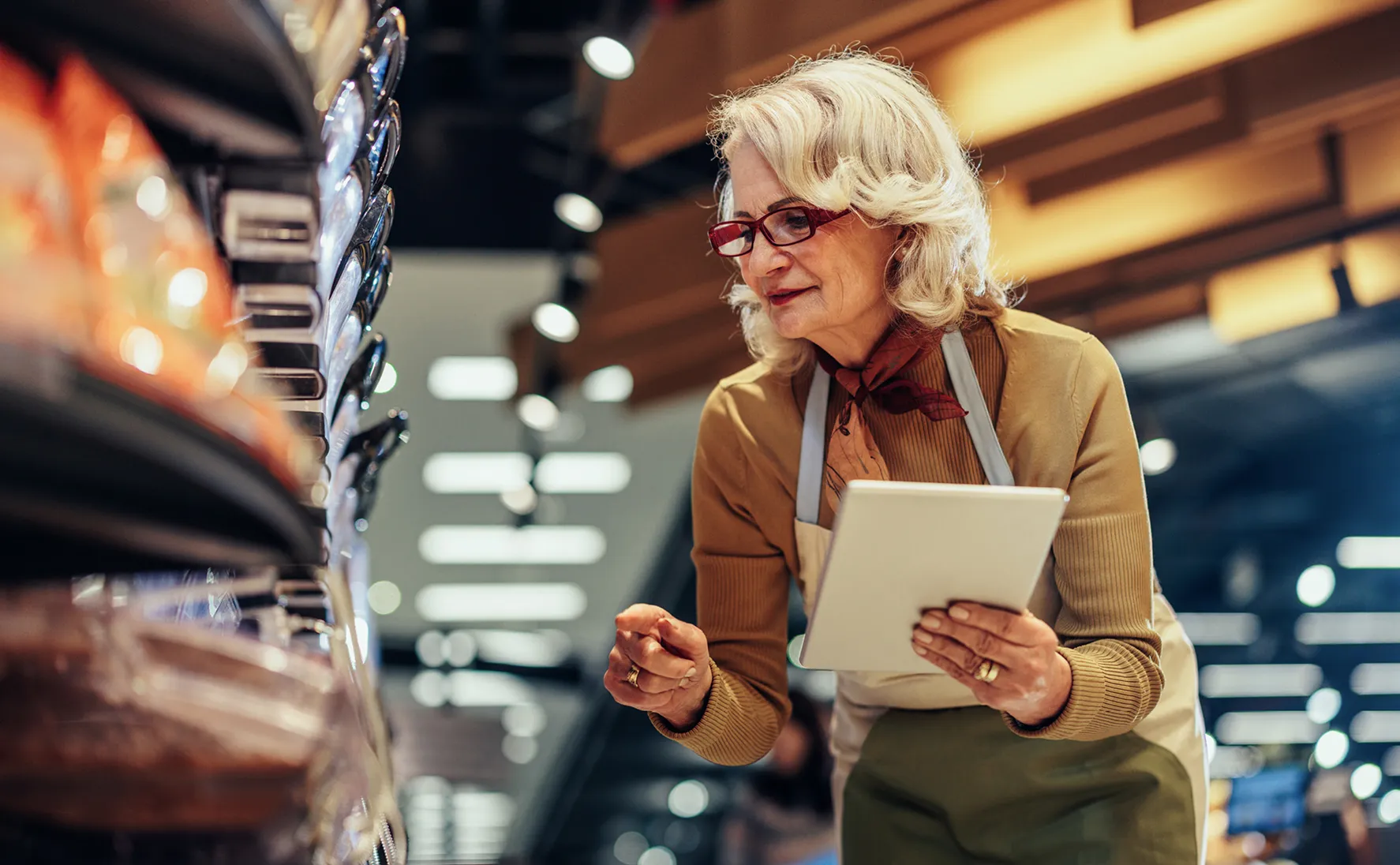 Older woman wearing glasses, a scarf, and a beige apron examining packaged meat in a grocery store while holding a digital tablet.