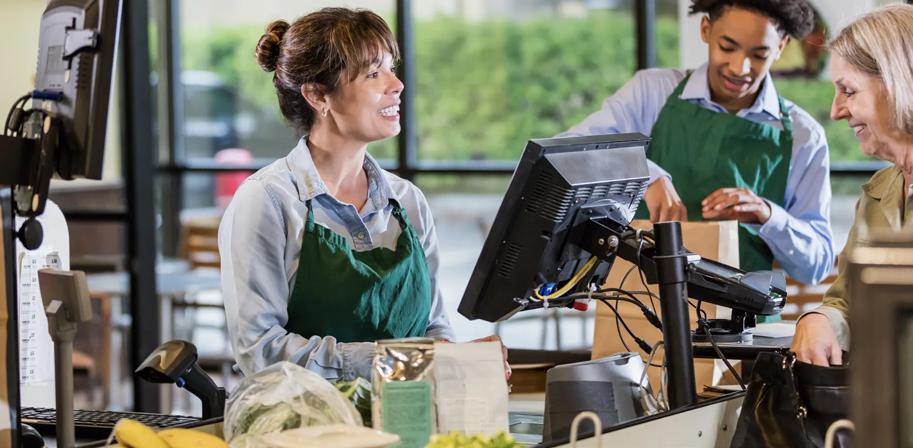 Smiling cashier in a green apron interacting with a customer at a grocery store checkout counter, while another employee in the background bags groceries.