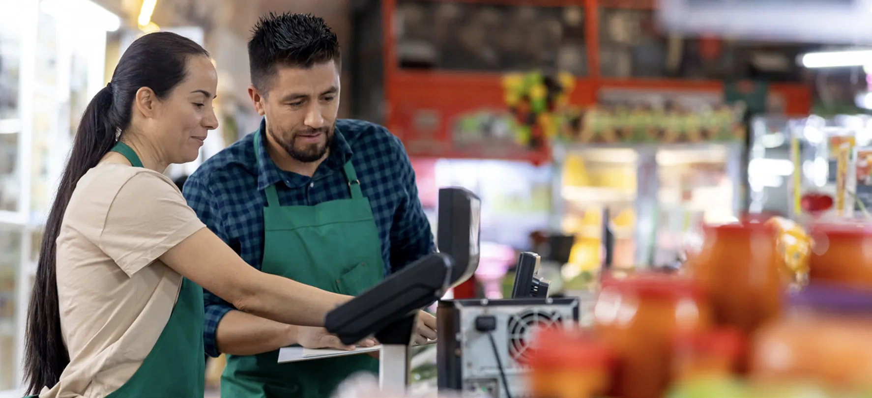 Two grocery store employees wearing green aprons are standing at a checkout station. The woman is pointing at the screen while the man observes, both appearing focused and engaged.