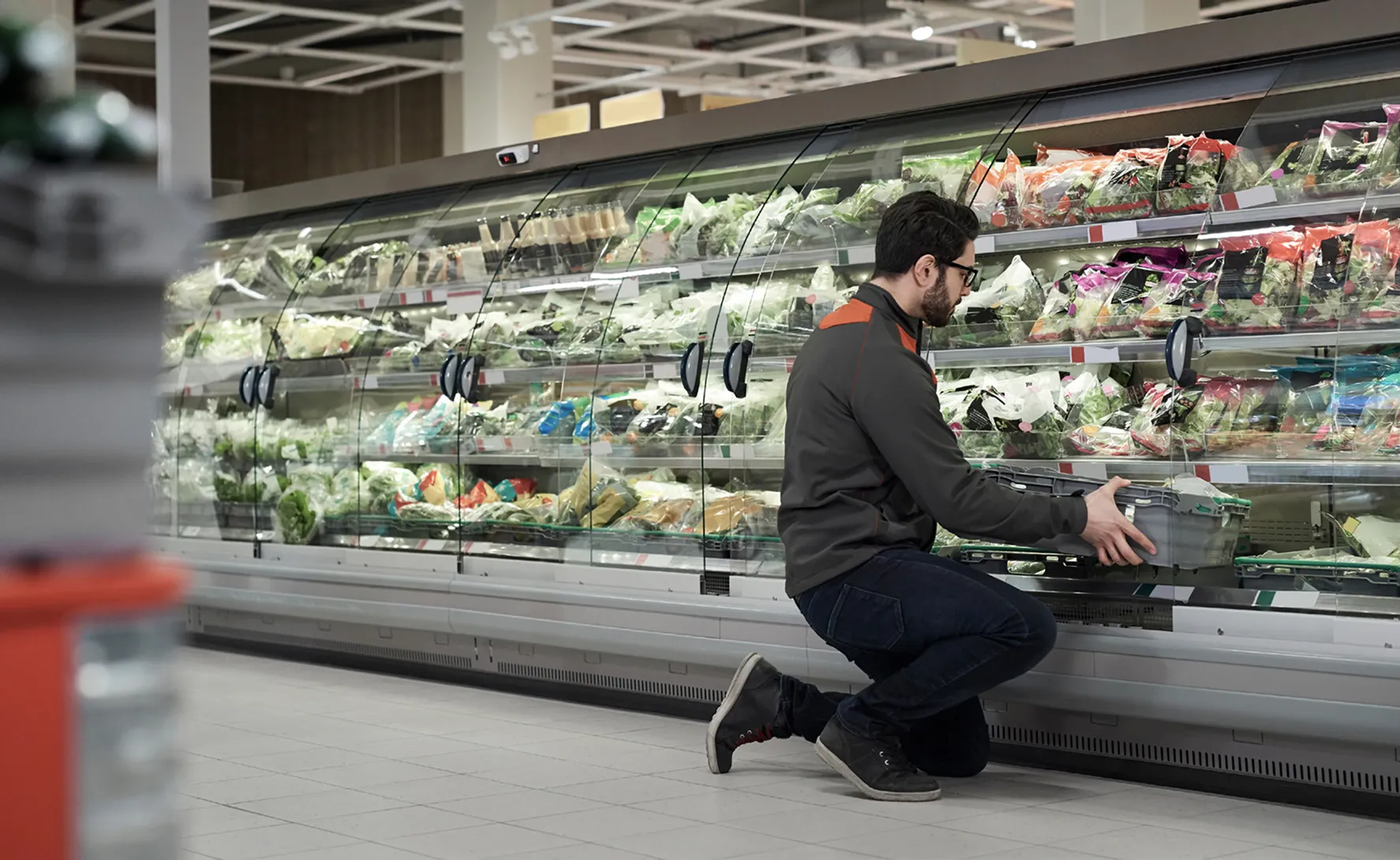 Man kneeling in front of a refrigerated display, restocking or organizing packaged salad greens in a grocery store.