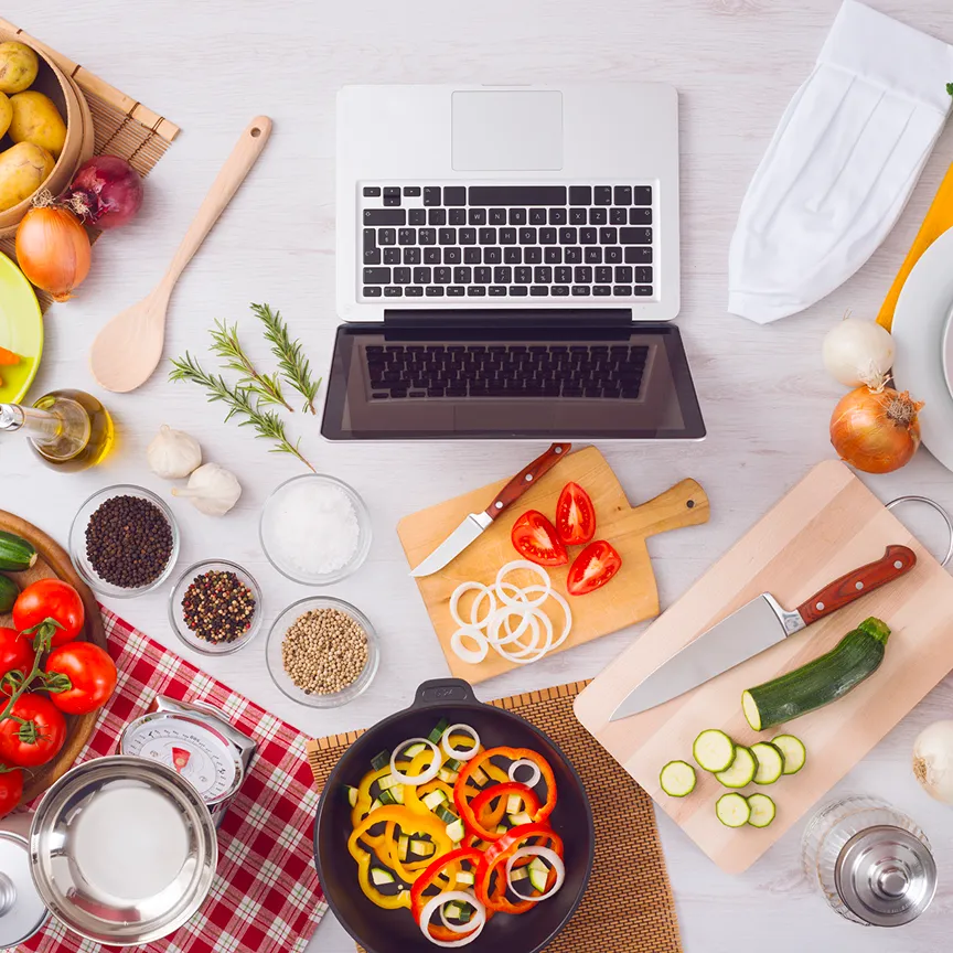 Overhead view of a cooking workspace featuring a laptop at the center, surrounded by fresh vegetables, spices, cutting boards, knives, a chef's hat, and kitchen utensils. Sliced ingredients like zucchini, bell peppers, onions, and tomatoes are arranged neatly, suggesting a on online recipe being followed.