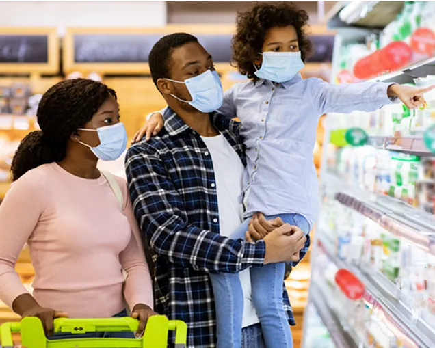 Family of three wearing face masks grocery shopping. The mother pushes a cart, the father holds a child who is pointing at items on a refrigerated store shelf.