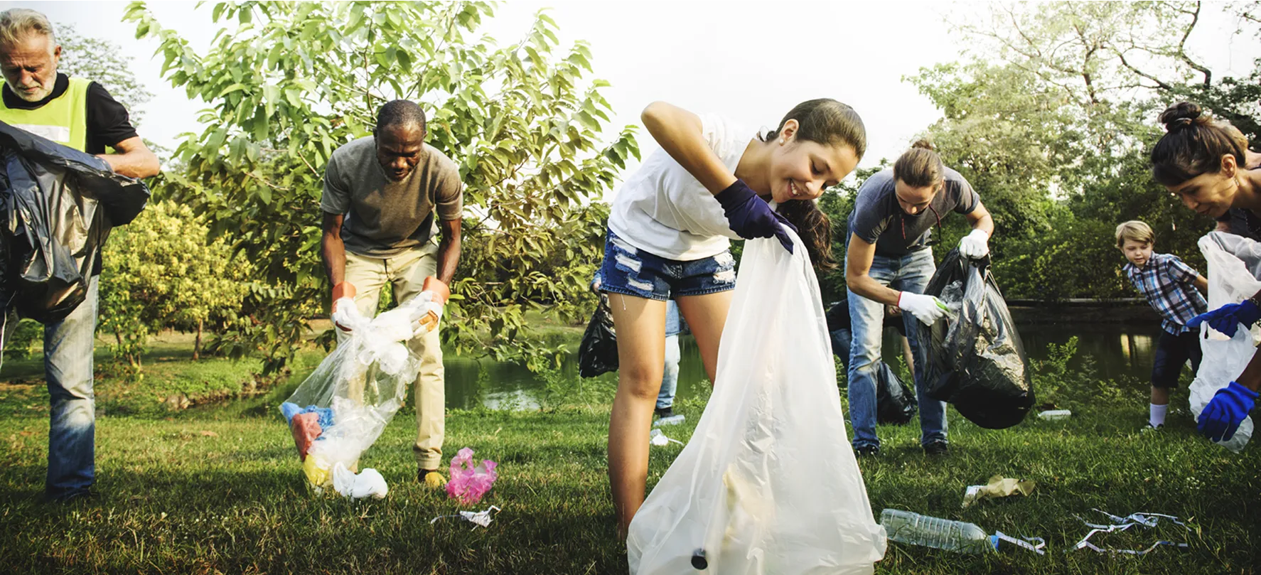 Group of people of various ages outdoors collecting litter in a grassy park area near a pond. They are wearing gloves and holding large trash bags while picking up plastic bottles and other debris.