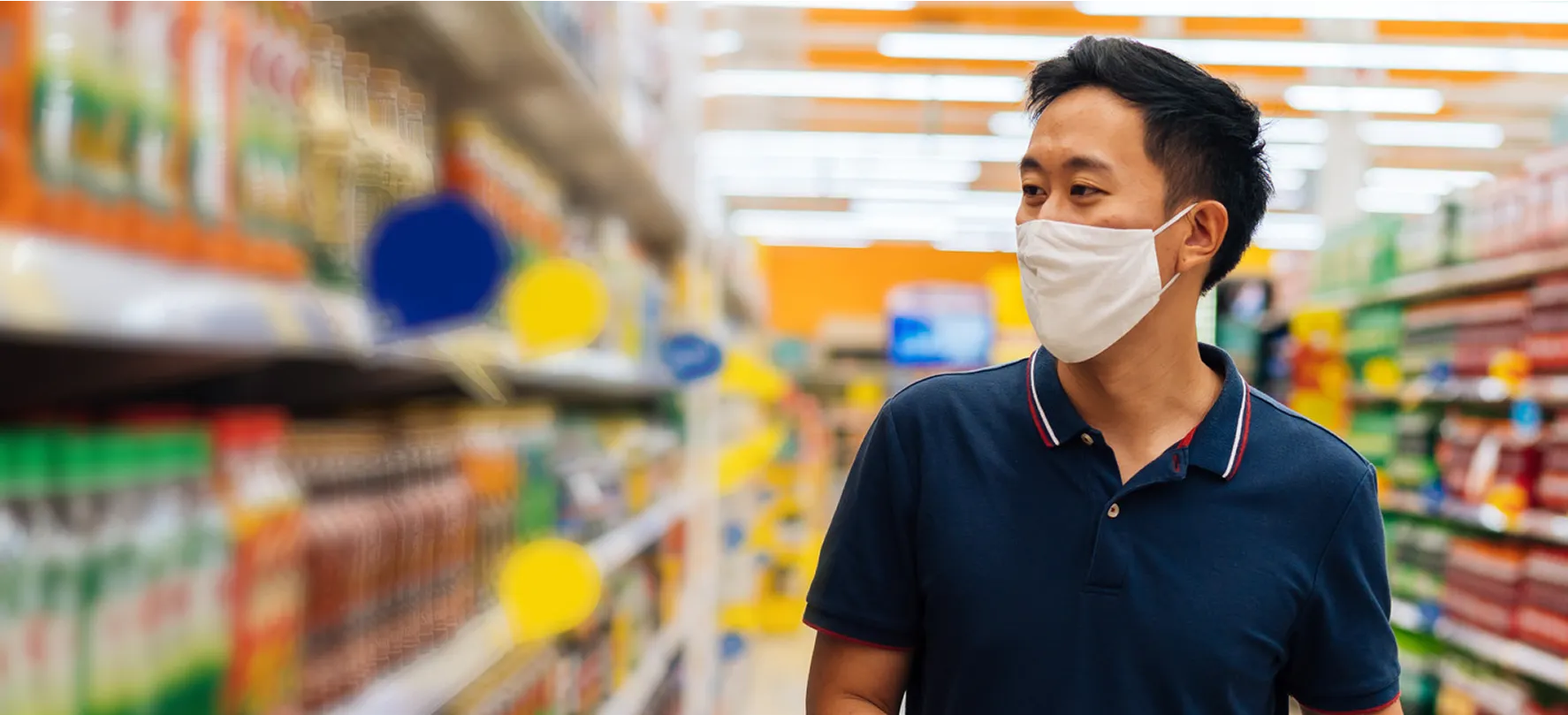 Man wearing a face mask walks through a brightly lit grocery store aisle lined with colorful bottled drinks.