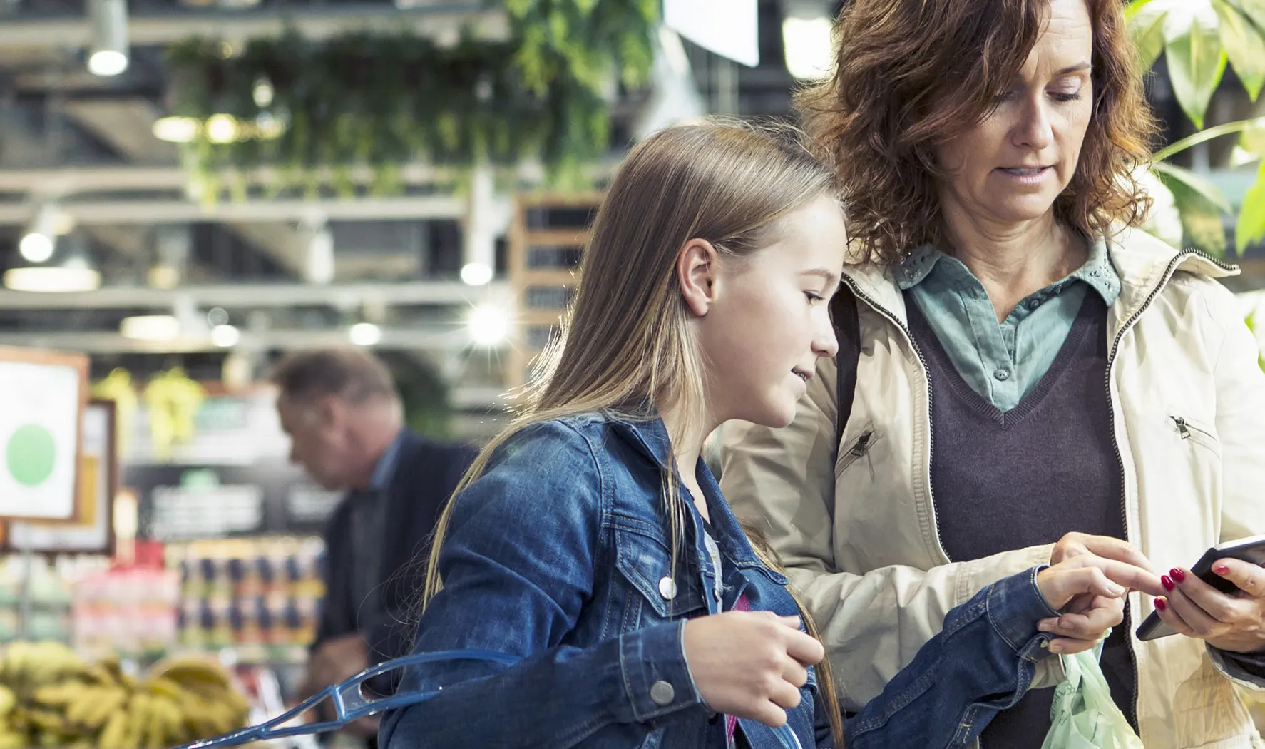 A woman and a young girl are grocery shopping together in a modern, well-lit supermarket. The woman is holding a smartphone and looking at it, while the girl points at the screen.