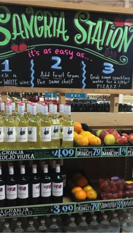 Trader Joe's "Sangria Station” retail display with a colorful chalkboard-style sign reading “It’s as easy as… 1. Pick a red or white wine, 2. Add fruit from the same shelf, 3. Grab some sparkling water for a little pizzazz!” Below the sign, shelves are stocked with bottles of white and red wine, fresh fruit like oranges, mangoes, and strawberries, and bottles of sparkling water, curated for building a DIY sangria.