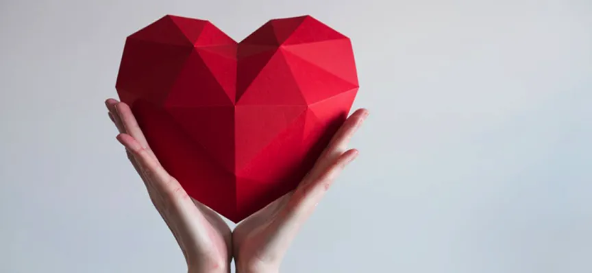 Hands holding a geometric, faceted red heart against a soft gray background.