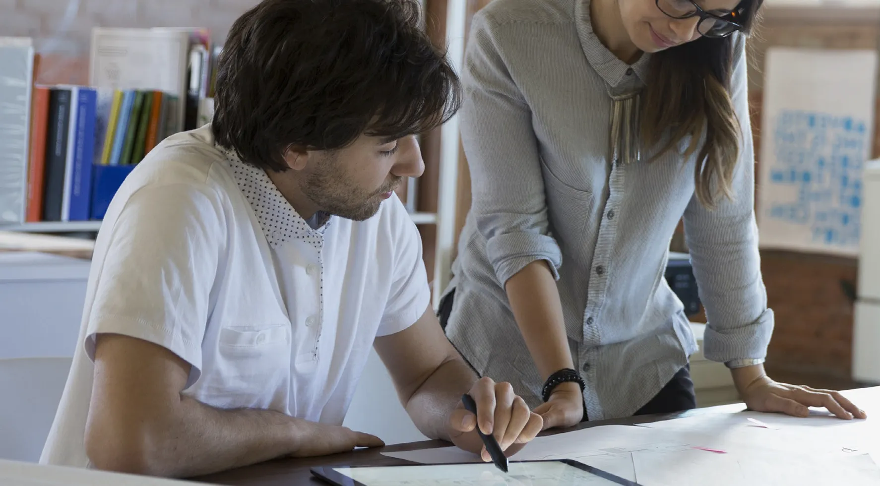 Two people collaborating at a desk in an office setting. A man is seated, using a stylus on a tablet, while a woman stands beside him, leaning over a large sheet of paper. They appear to be working on a design or planning project, with shelves of books and office supplies in the background.